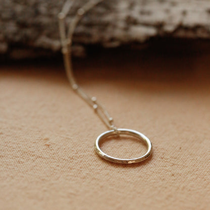 A ring of sterling silver sits on a satellite chain with a background of sand coloured natural cotton tablecloth. A piece of bark is visible in the background, faded out.