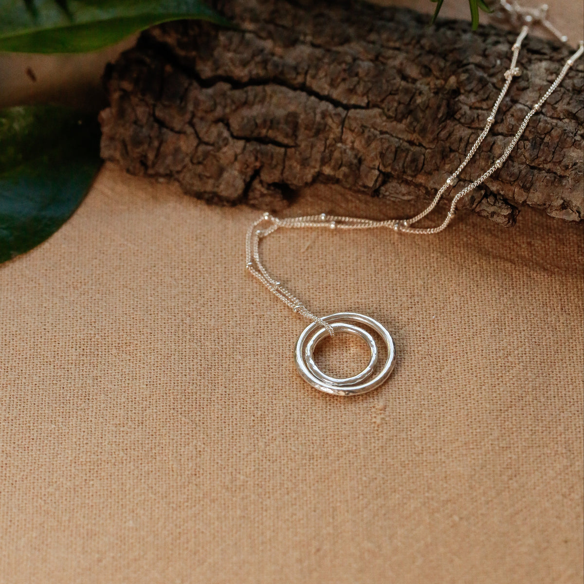 A silver double-ring pendant necklace on a delicate chain, styled on a textured beige surface with a piece of rustic wood and green leaves in the background.