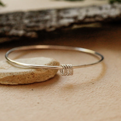 A thin silver bangle bracelet with a textured spiral wrap detail, resting on a small stone over a beige linen surface with a blurred bark background.