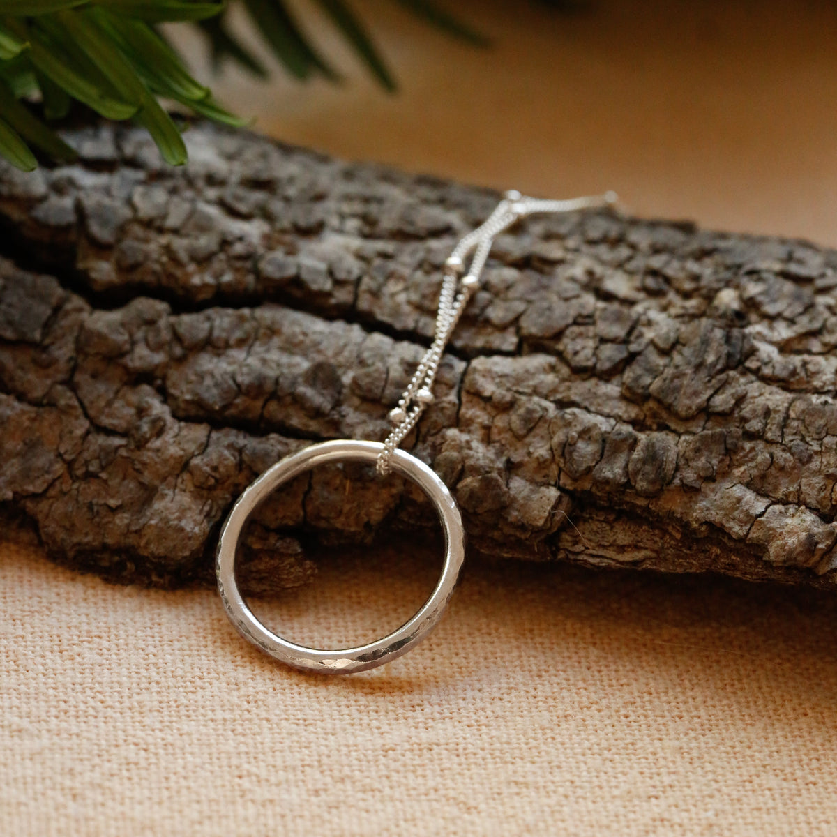A silver ring pendant necklace resting on a piece of textured bark, with pine needles in the background on a beige fabric surface.