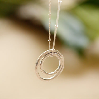 A silver necklace with a double-ring pendant and beaded chain, shown hanging against a softly blurred warm beige and green background.