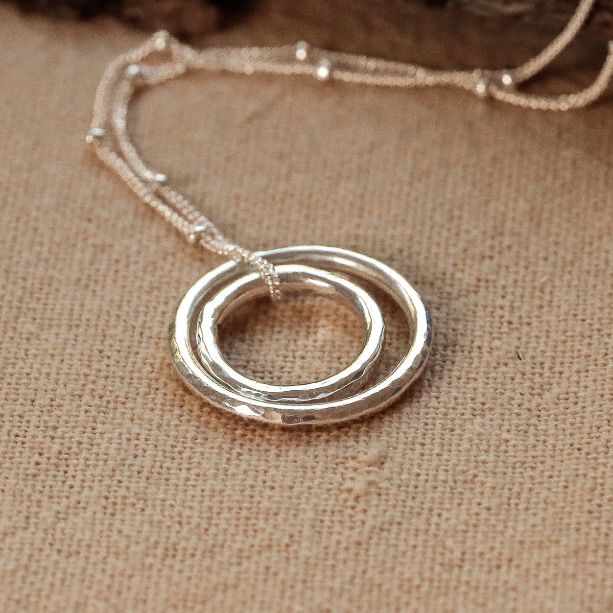 A silver necklace with a double-ring pendant resting on a textured beige fabric surface next to a piece of bark and green pine needles.