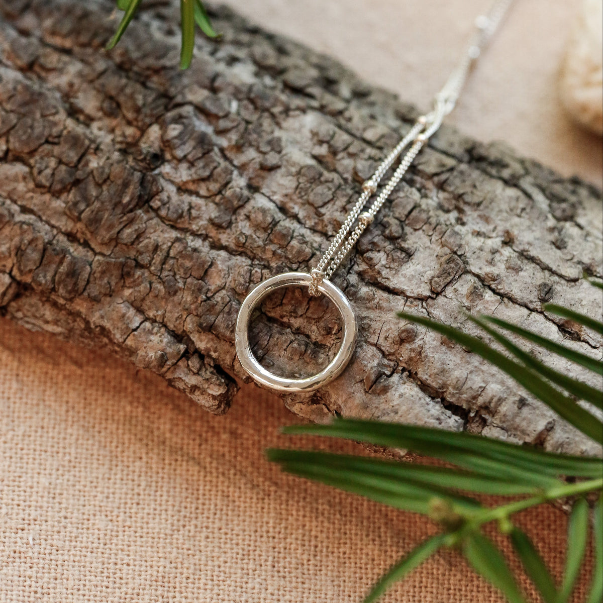 A silver pendant necklace with a simple circular ring on a delicate chain, styled on a piece of textured bark with green pine branches on a beige fabric background.