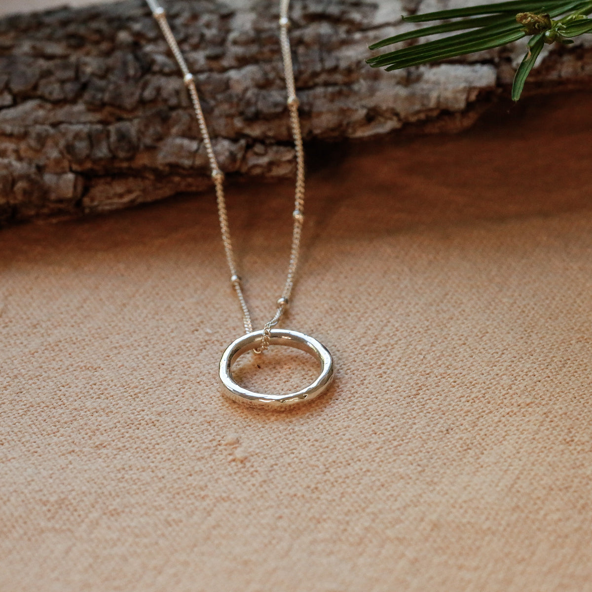 A small sterling silver open circle pendant necklace on a delicate satellite chain, displayed on a textured beige surface with a piece of bark and green pine needles in the background.