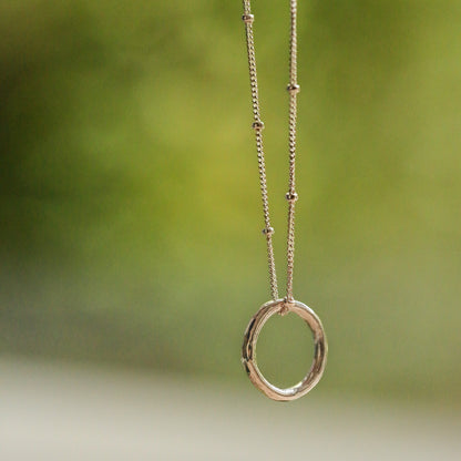 A delicate silver necklace with a small open ring pendant and tiny bead accents on the chain, shown against a soft, blurred green and white outdoor background.