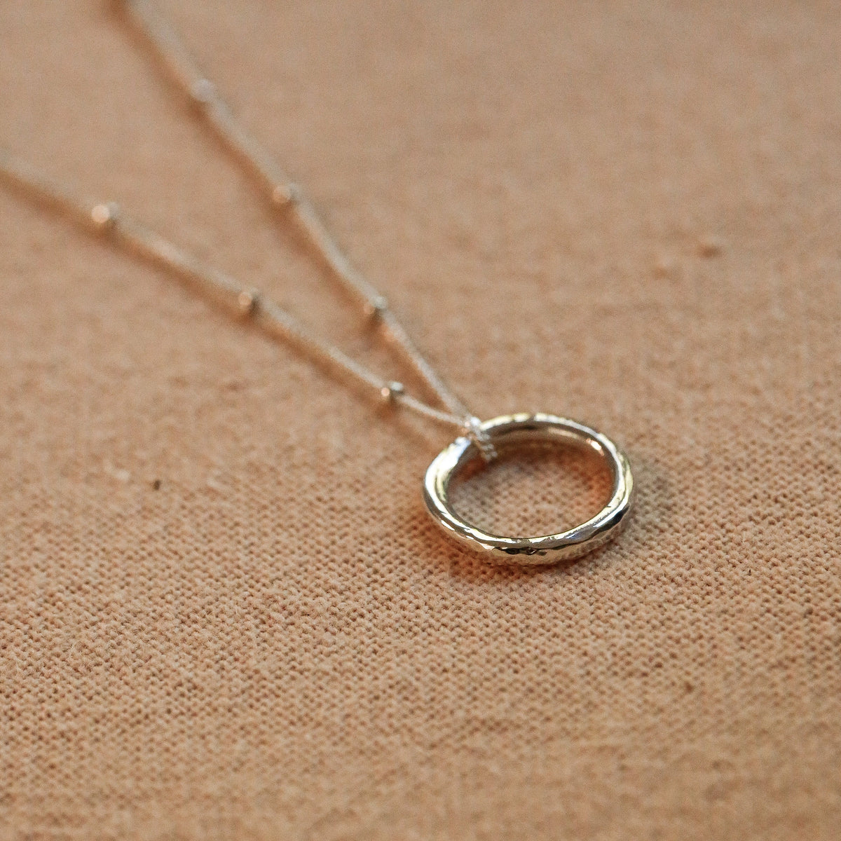A silver open ring pendant necklace on a delicate chain, displayed on a textured tan fabric background.