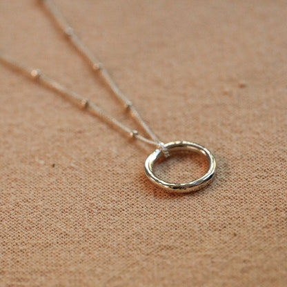A silver open ring pendant necklace on a delicate chain, displayed on a textured tan fabric background.