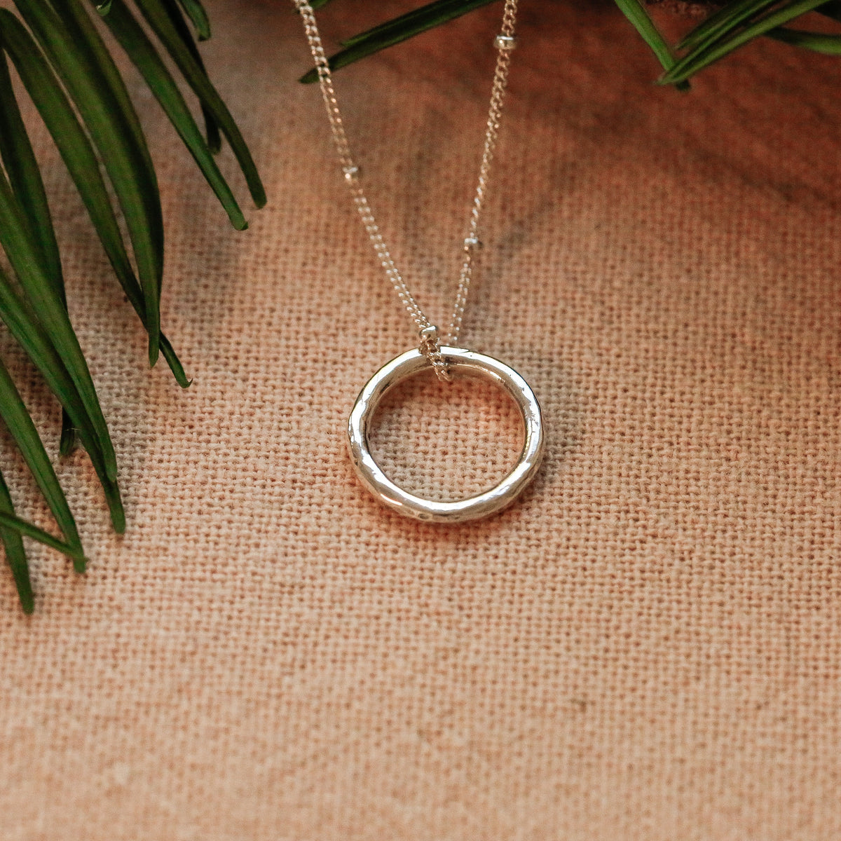 A silver ring pendant necklace on a delicate chain, displayed on a textured beige fabric background with green leaves in the corner.