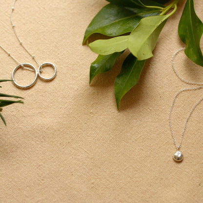 Three silver necklaces with minimalist pendants are arranged on a textured beige fabric background with green leaves as styling props. One necklace features two interlocking rings, while the other two have small polished silver beads.