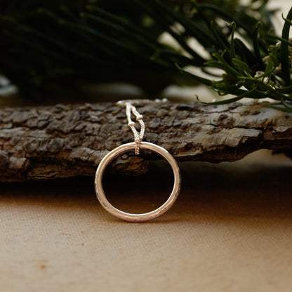 A sterling silver open circle pendant with a small loop at the top, resting on a piece of textured bark with green foliage blurred in the background.