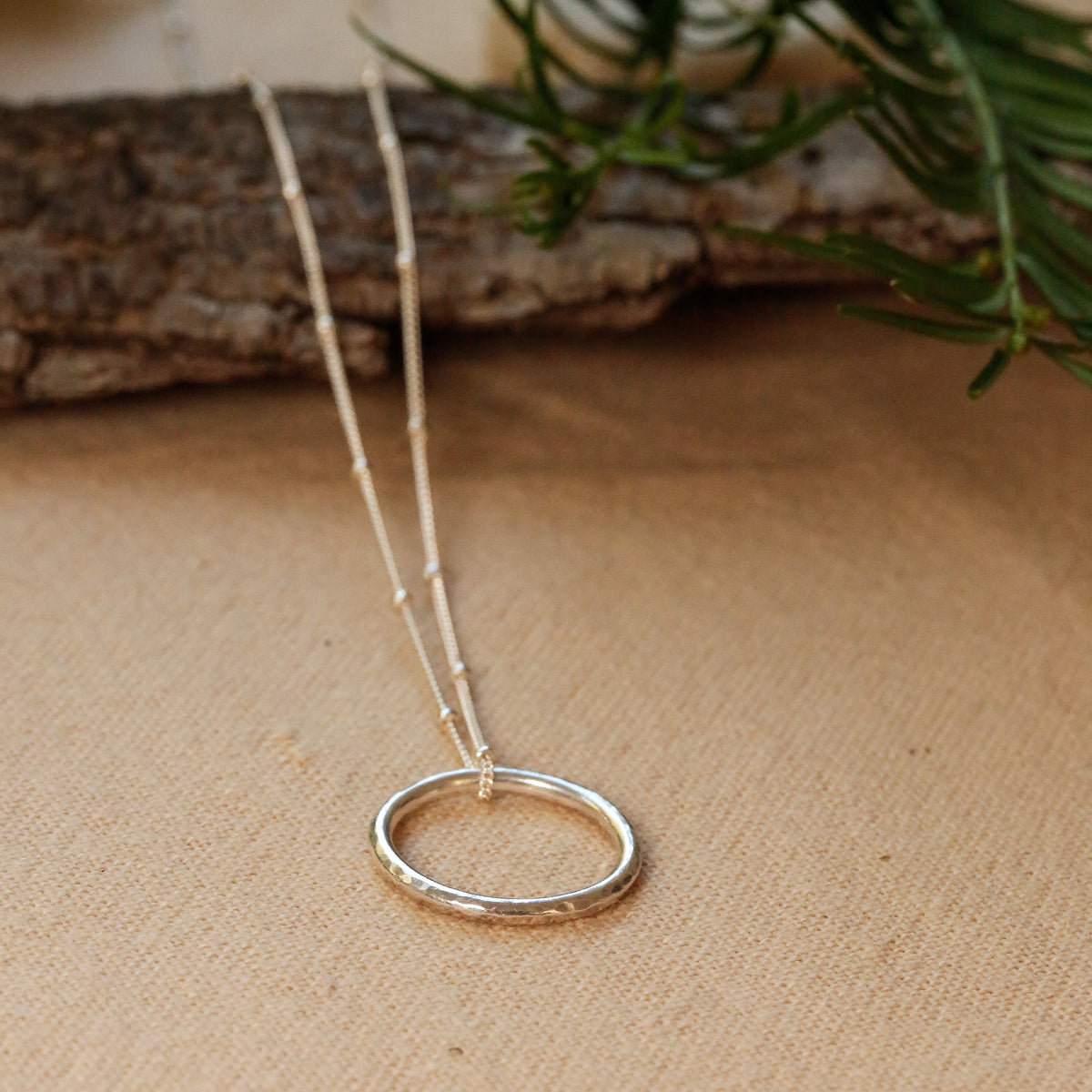 A delicate silver necklace with a textured circular pendant, resting on a beige linen surface with a rustic wood branch and green pine needles in the background.