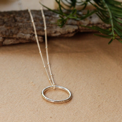 A delicate silver necklace with a textured circular pendant, resting on a beige linen surface with a rustic wood branch and green pine needles in the background.