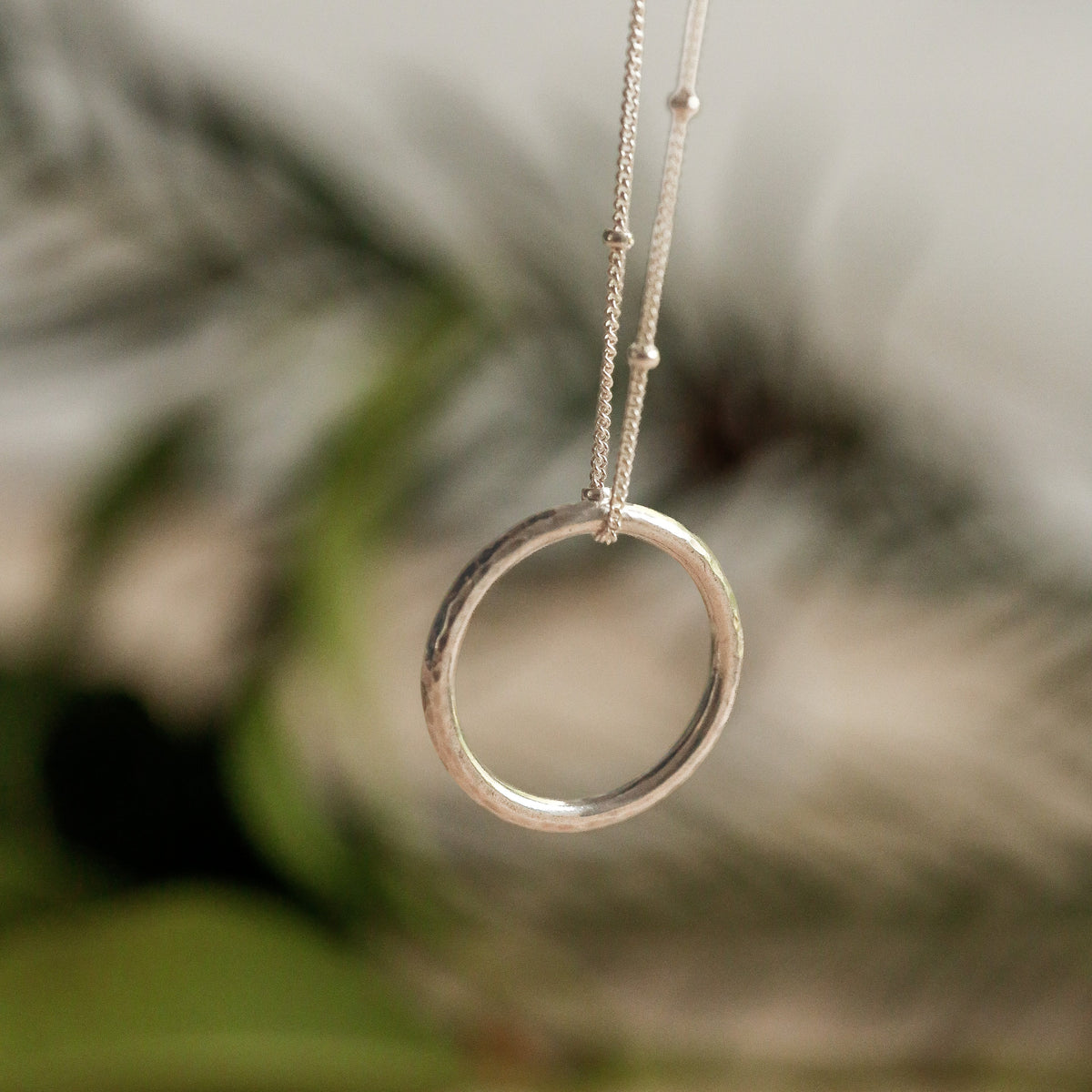 A sterling silver silver minimalist ring pendant necklace hanging from a delicate chain, set against a soft-focus background of green leaves and beige sand.