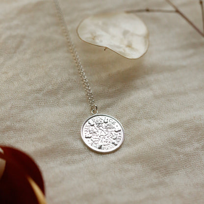 A silver acorns coin pendant necklace with an engraved circular medallion, resting on a textured beige linen fabric with dried botanical elements in the background.