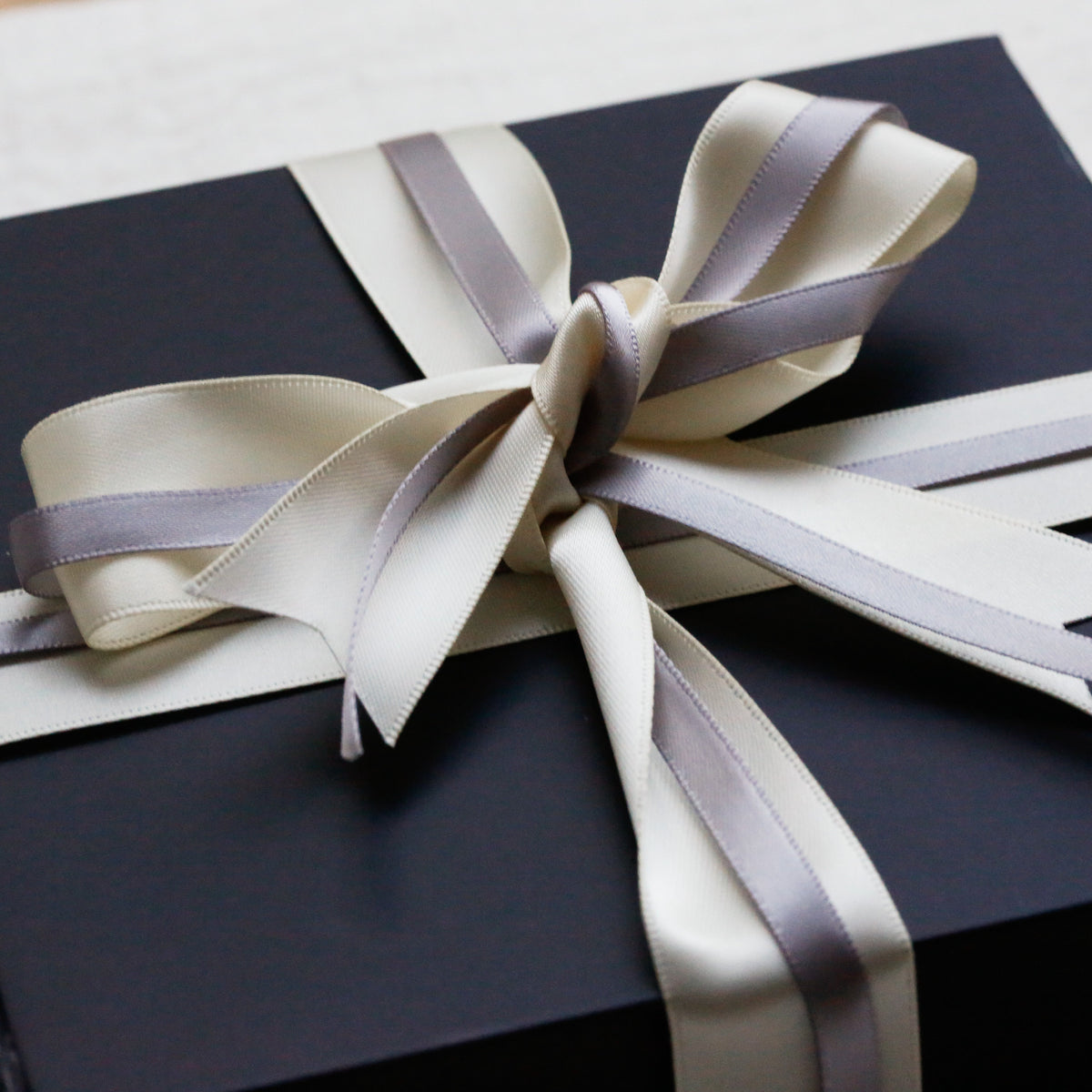 A square black gift box wrapped with a cream satin ribbon and bow, photographed close up on a light neutral surface.