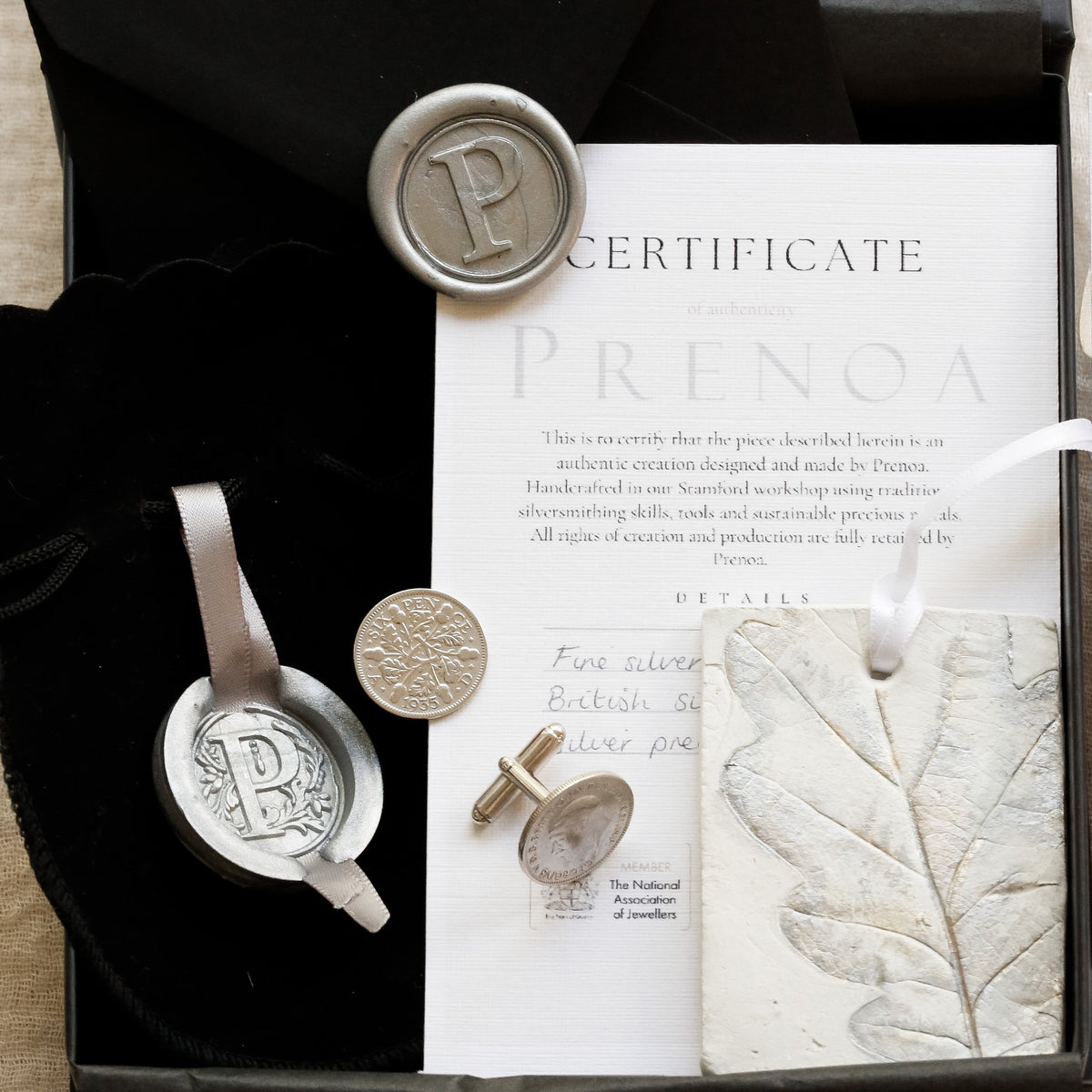 A luxury gift set from Acorns & Oaks featuring a silver cufflinks with a wax seal, a black velvet pouch, a certificate, and a wax seal stamp inside a black presentation box.