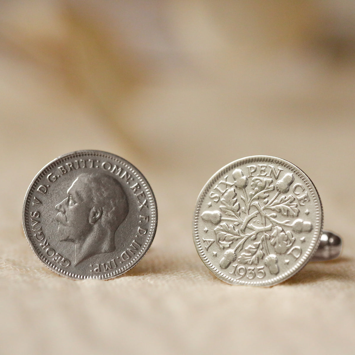 A pair of silver cufflinks shaped like vintage British half crowns, displayed on a soft beige textured surface.