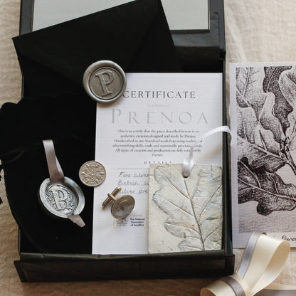 A styled flat-lay of a gift set on a beige fabric background, featuring a black presentation box with a wax seal, a black velvet pouch, silver cufflinks a certificate card, a leaf-printed tag, and an oak leaf illustration print.