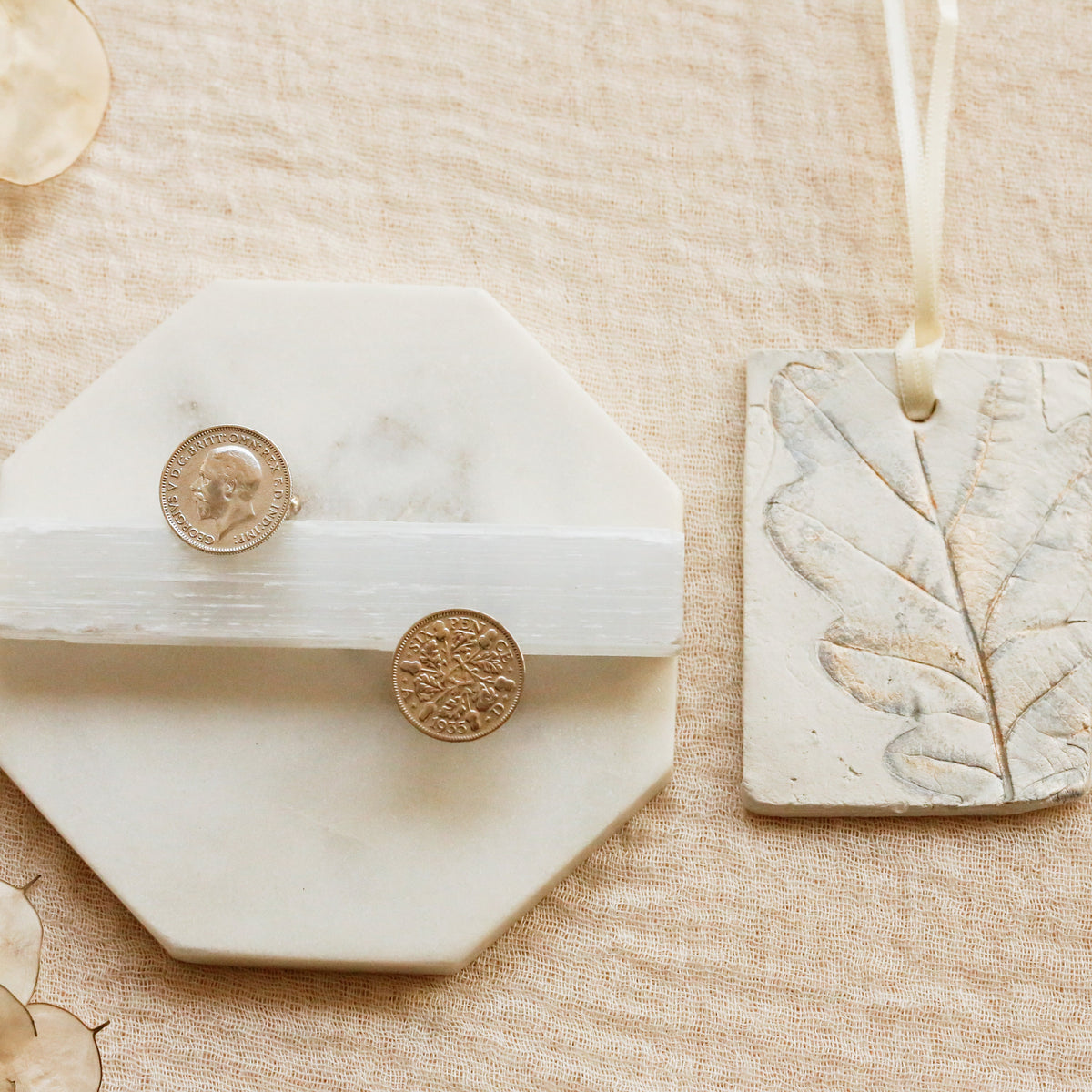 A styled flat-lay of a white octagonal marble ring holder with a white band holding two silver sixpence coin cufflinks, next to a rectangular stone tag with an embossed leaf design hanging from a ribbon, all on a beige linen background.