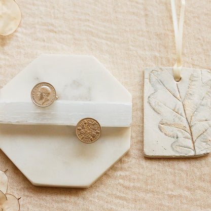 A styled flat-lay of a white octagonal marble ring holder with a white band holding two silver sixpence coin cufflinks, next to a rectangular stone tag with an embossed leaf design hanging from a ribbon, all on a beige linen background.
