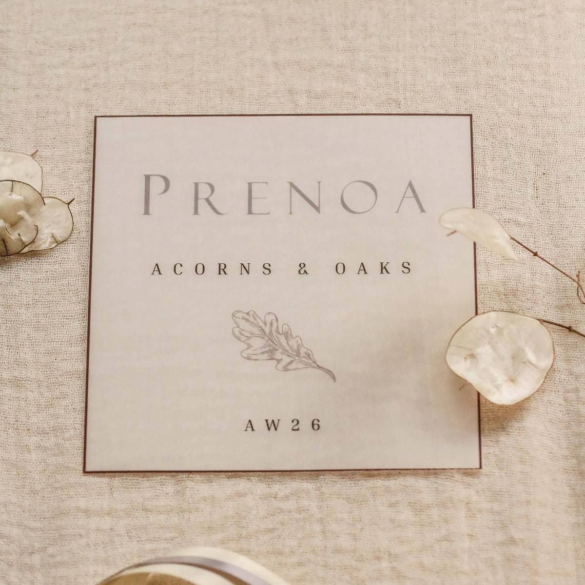 A square cream wedding card with elegant gray typography reading 'PRENOA', 'ACORNS & OAKS', and 'AW26', styled on a textured beige linen background with dried flowers and a cream ribbon.
