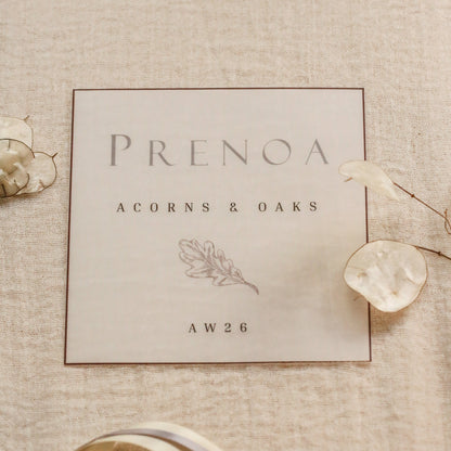 A square cream wedding card with elegant gray typography reading 'PRENOA', 'ACORNS & OAKS', and 'AW26', styled on a textured beige linen background with dried flowers and a cream ribbon.