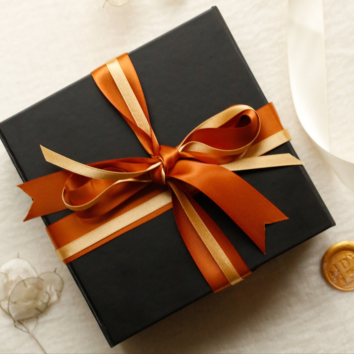 A black gift box wrapped with a wide orange and gold ribbon, tied in a bow, photographed on a light textured surface with decorative dried flowers and a white ribbon roll nearby.