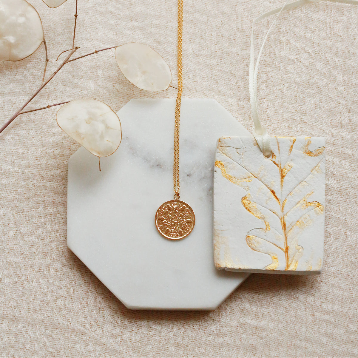 A gold coin pendant necklace displayed on a white marble octagonal tray, next to a white ceramic tag with gold leaf botanical patterns, on a textured beige fabric background with dried eucalyptus leaves.