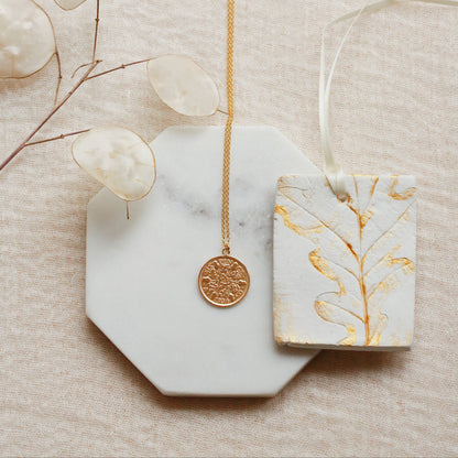 A gold coin pendant necklace displayed on a white marble octagonal tray, next to a white ceramic tag with gold leaf botanical patterns, on a textured beige fabric background with dried eucalyptus leaves.