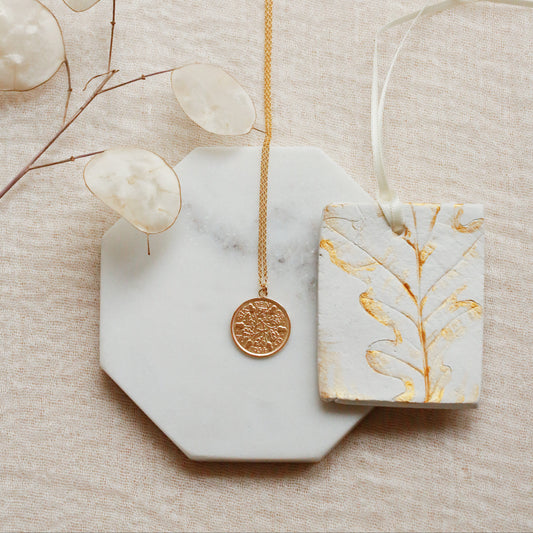 A gold coin pendant necklace displayed on a white marble octagonal tray, next to a white ceramic tag with gold leaf botanical patterns, on a textured beige fabric background with dried eucalyptus leaves.