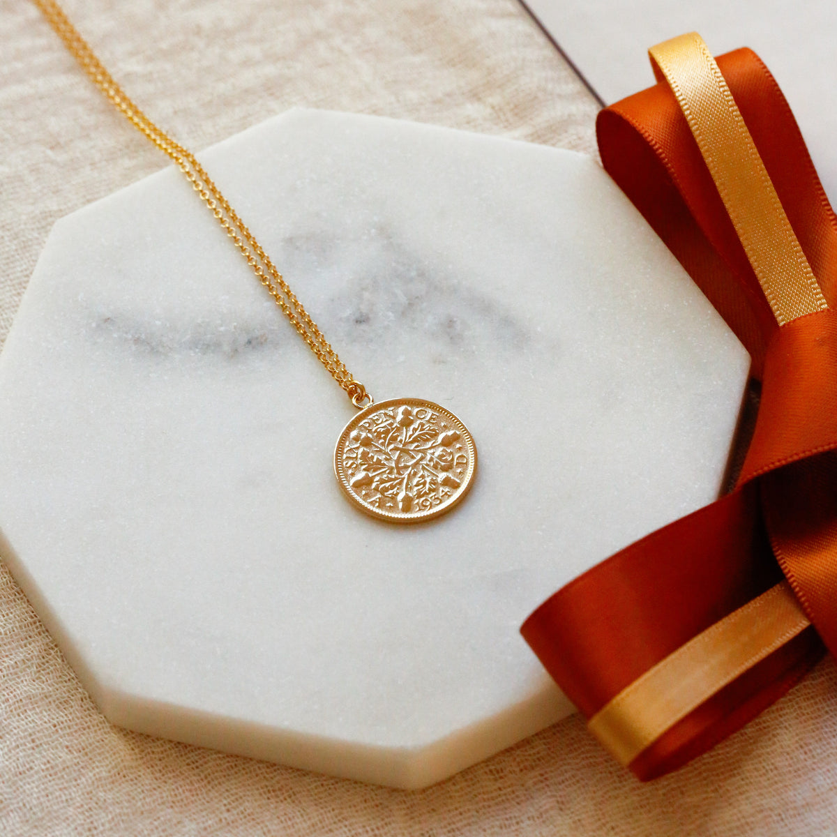 A gold coin necklace with an round sixpence coin, acorns and oak leaves pendant and gold vermeil chain, displayed on a white marble octagonal platform with a burnt orange ribbon and a beige fabric background.