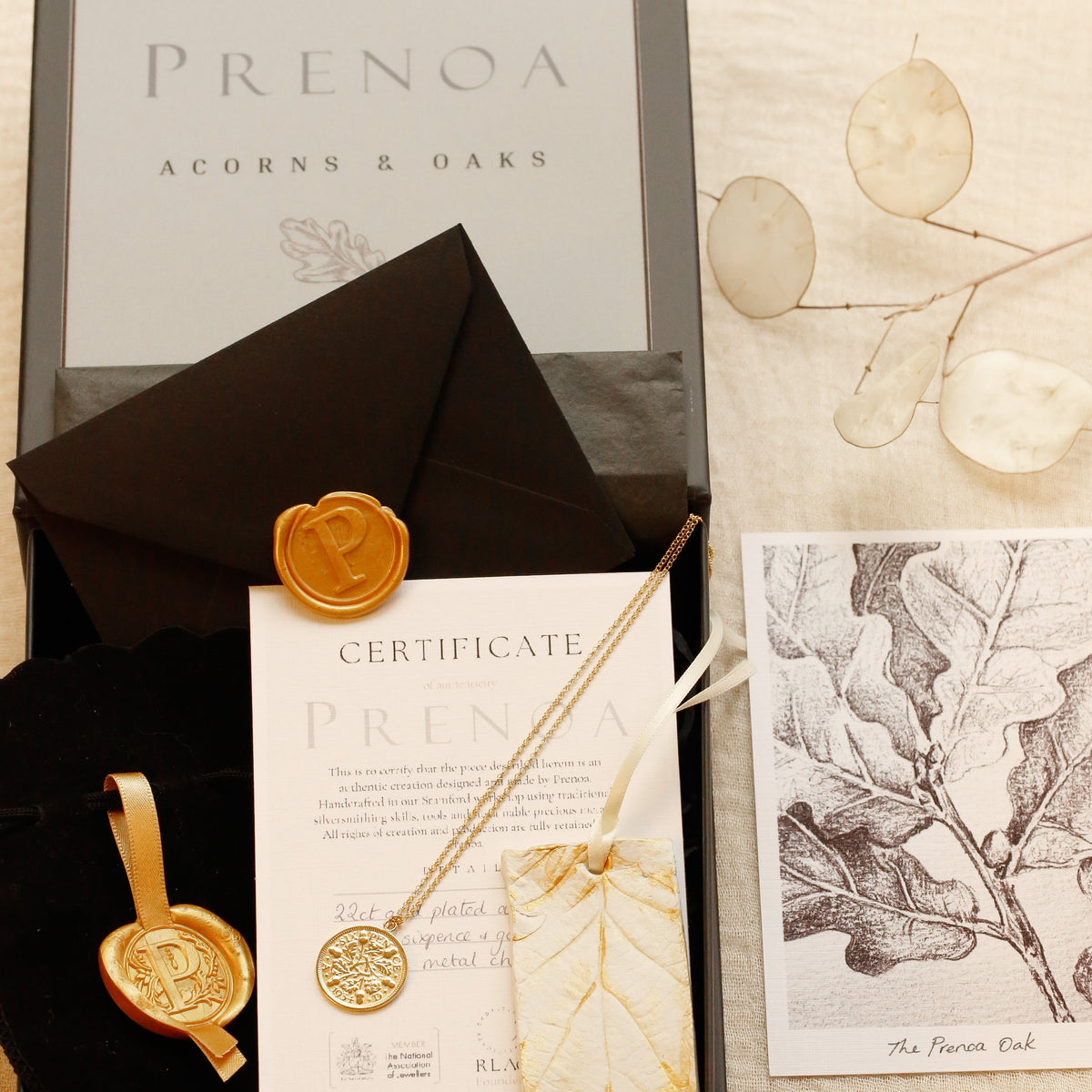 A boxed jewelry gift set from Prenoa Acorns & Oaks arranged on a light fabric background. The set includes a black presentation box, a black envelope, a gold wax seal, a certificate card, a leaf-shaped pendant necklace, and a botanical print card.