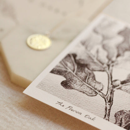 A small round gold pendant necklace displayed on a cream-colored card next to a botanical print labeled 'The Prenoa Oak', set on a textured beige fabric surface.