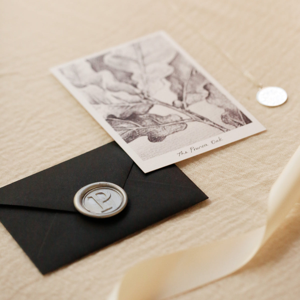 A styled flat-lay of a black envelope with a silver wax seal, next to a botanical illustration card reading 'The Prenoa Oak' on a textured beige background with satin cream ribbon.