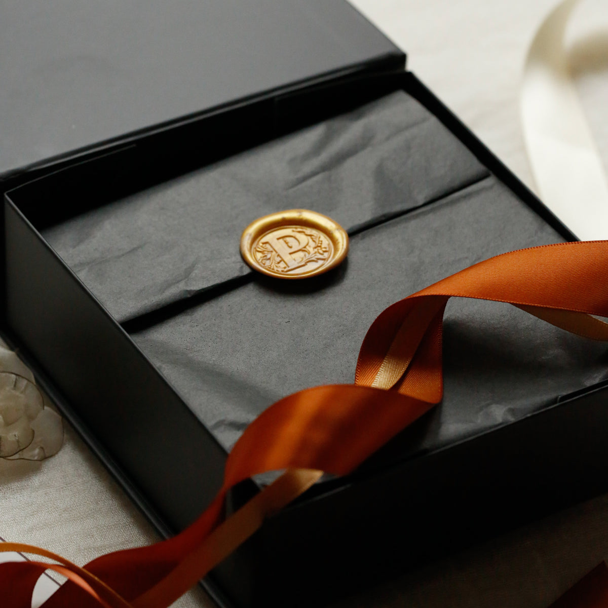 A round gold wax seal with an embossed monogram 'P' resting inside an open black presentation box lined with black tissue paper, with an orange ribbon draped across the front.