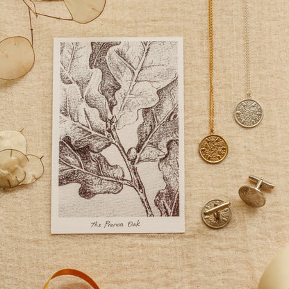 A styled flat-lay featuring a square art print titled 'The Prenoa Oak' with a black and white oak leaf illustration. The print is displayed on a textured beige linen background alongside gold and silver jewelry, including a gold chain necklace, a silver pendant, and a silver cufflink, along with dried botanical elements and ribbon.