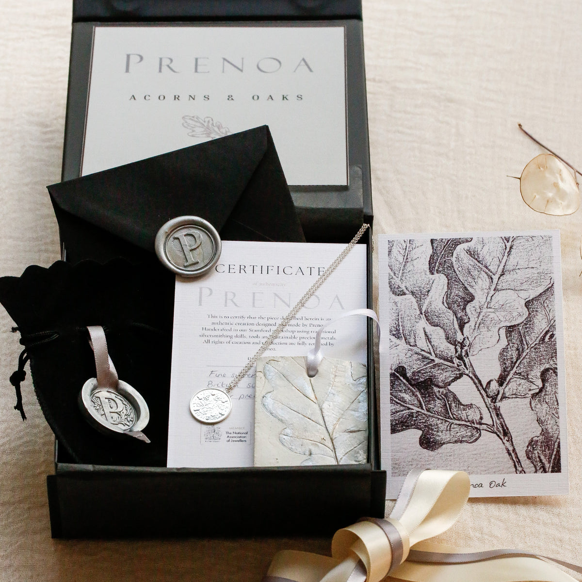 A luxury gift set from Prenoa Acorns & Oaks, featuring a silver oak leaf pendant necklace, a silver wax seal, a certificate, and a botanical print card, all presented in a black gift box with a cream ribbon on a beige linen background.