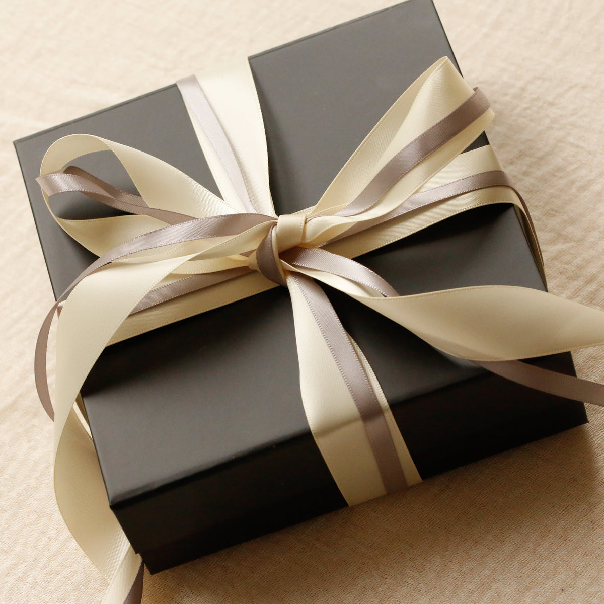 A black gift box wrapped with a cream and taupe satin ribbon, tied in a bow, on a textured beige fabric background.