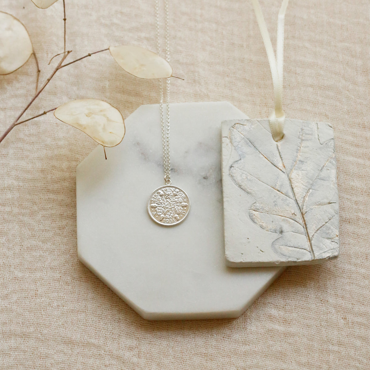 A silver acorns and oaks coin pendant necklace  and a rectangular silver leaf charm on a cream ribbon, displayed on a white marble octagonal tray over a beige linen background.