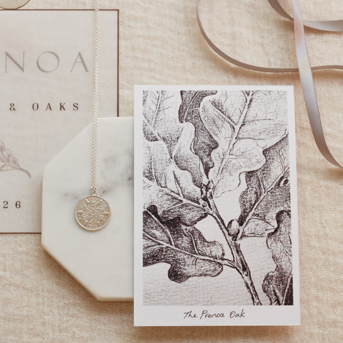 A styled flat-lay featuring a custom monochrome oak leaf art print card titled 'The Prenoa Oak', displayed alongside a silver coin pendant on a white marble hexagonal tray and a gray ribbon on a beige linen background.