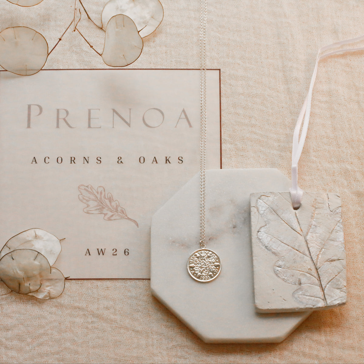 A styled flat-lay of jewelry and packaging on a textured beige fabric background. The set includes a silver-toned pendant necklace on a white octagonal display block, a rectangular tag with an embossed leaf design, a branded card reading 'PRENOA ACORNS & OAKS', and decorative dried botanical elements.