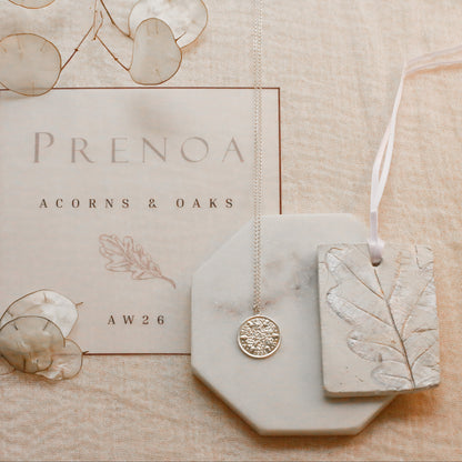 A styled flat-lay of jewelry and packaging on a textured beige fabric background. The set includes a silver-toned pendant necklace on a white octagonal display block, a rectangular tag with an embossed leaf design, a branded card reading 'PRENOA ACORNS & OAKS', and decorative dried botanical elements.