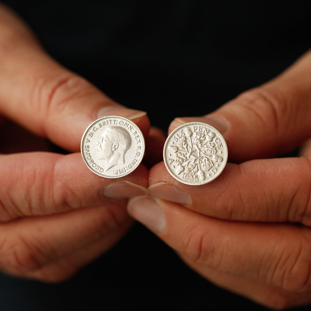 A close-up of two hands holding a pair of silver coin cufflinks against a dark background.
