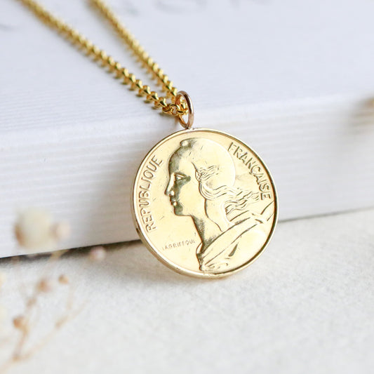 Marianne French gold necklace with a coin pendant on a white surface with dried flowers.