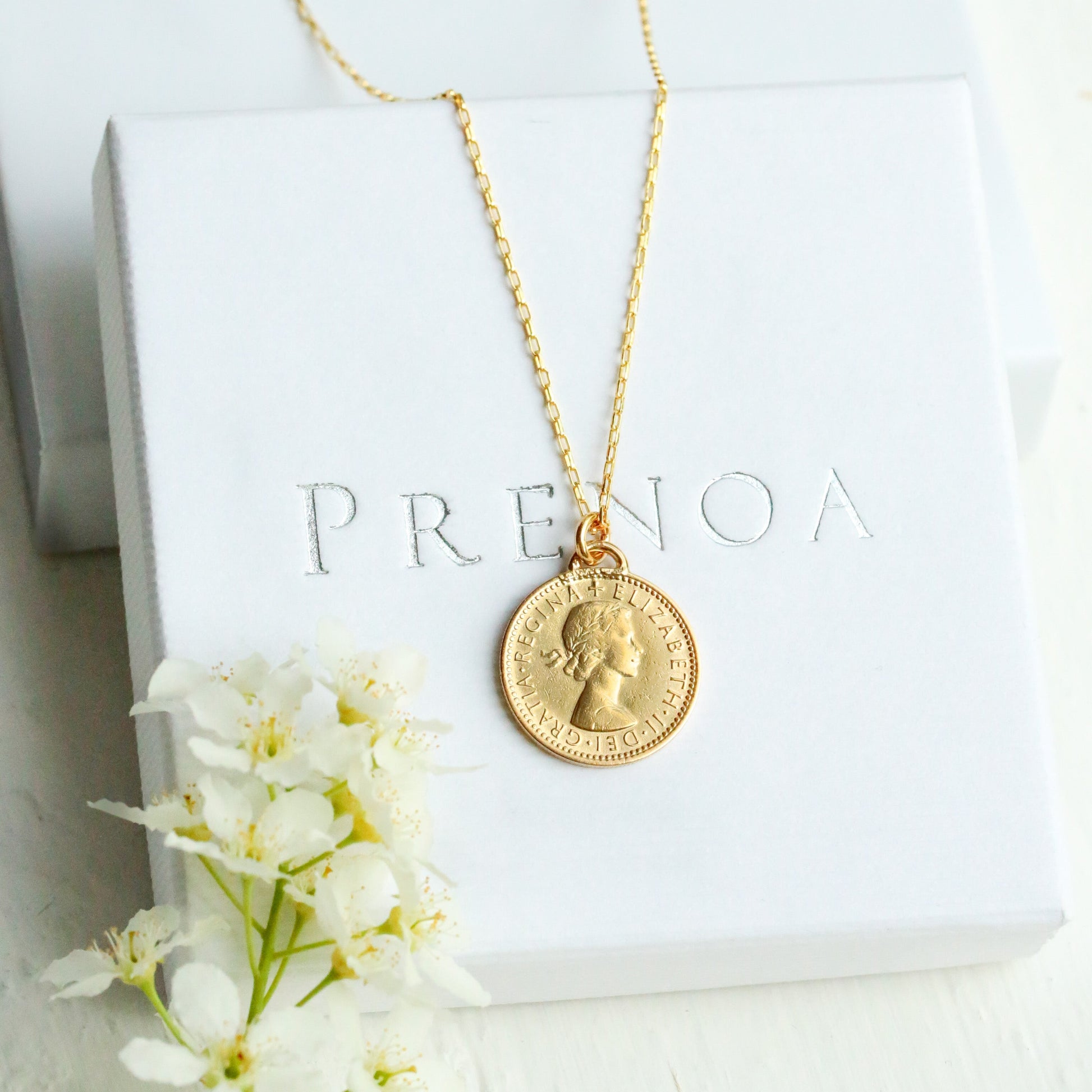 Gold sixpence necklace with a pendant on a white box with 'PRENOA' branding, surrounded by white flowers.