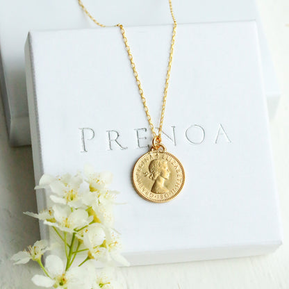 Gold sixpence necklace with a pendant on a white box with 'PRENOA' branding, surrounded by white flowers.