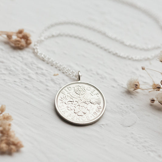 Silver sixpence coin necklace with a coin pendant on a light wooden surface with dried flowers.