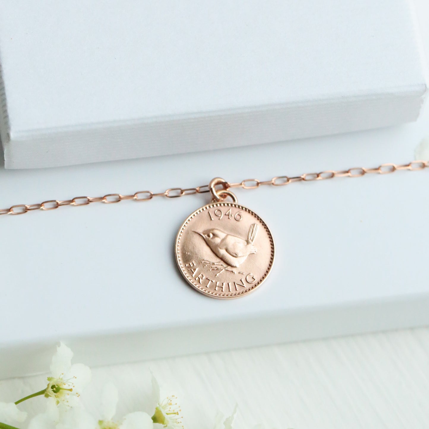 Rose gold necklace with a coin pendant on a white box with 'PRENOT' branding, surrounded by white flowers.