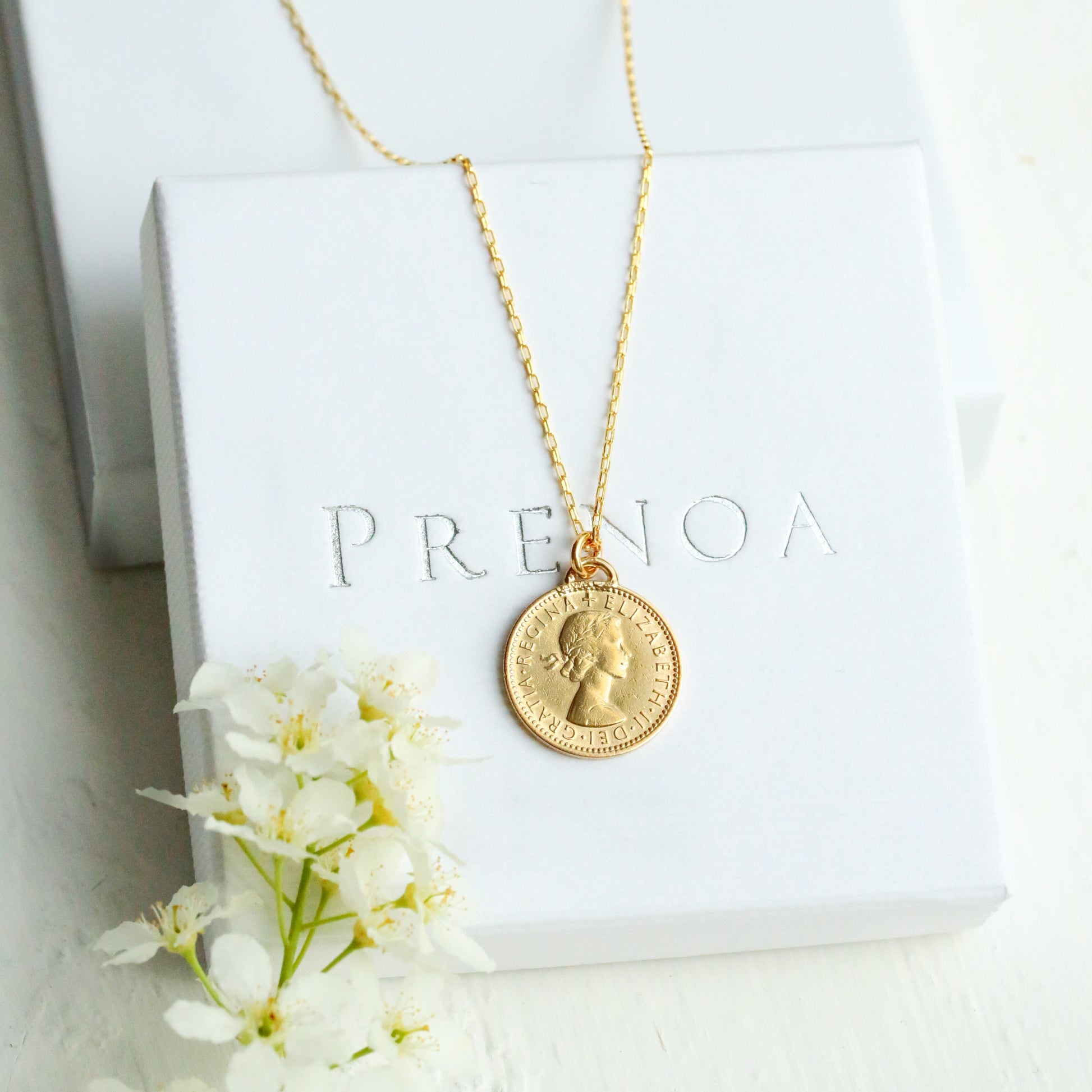 Gold necklace with a pendant on a white box with 'PRENOA' branding, surrounded by white flowers.