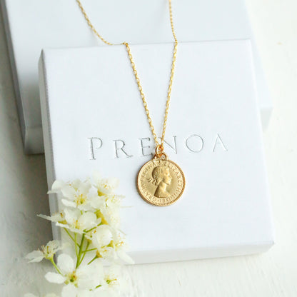 Gold necklace with a pendant on a white box with 'PRENOA' branding, surrounded by white flowers.